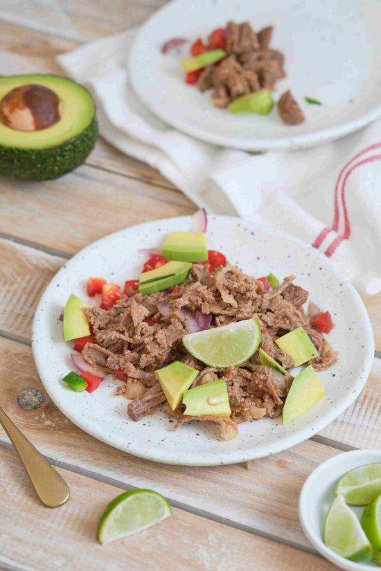 Two plates of Beef Carnitas are placed on a wooden surface, with slices of lemon and avocado.