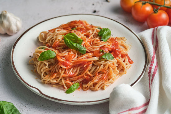 A plate of spaghetti marinara on a marble white surface with some tomatoes, garlic and a kitchen cloth.