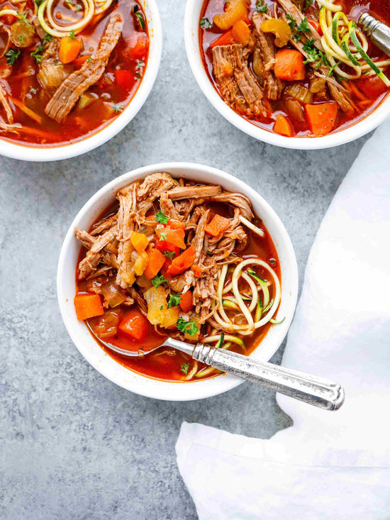 Three bowls of Smoky Brisket Zoodle Soup on a grey plain surface.