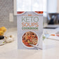 The Keto Soups Cookbook stands on a plain marble surface. Flower pots, vegetables, and some bottles can be seen.