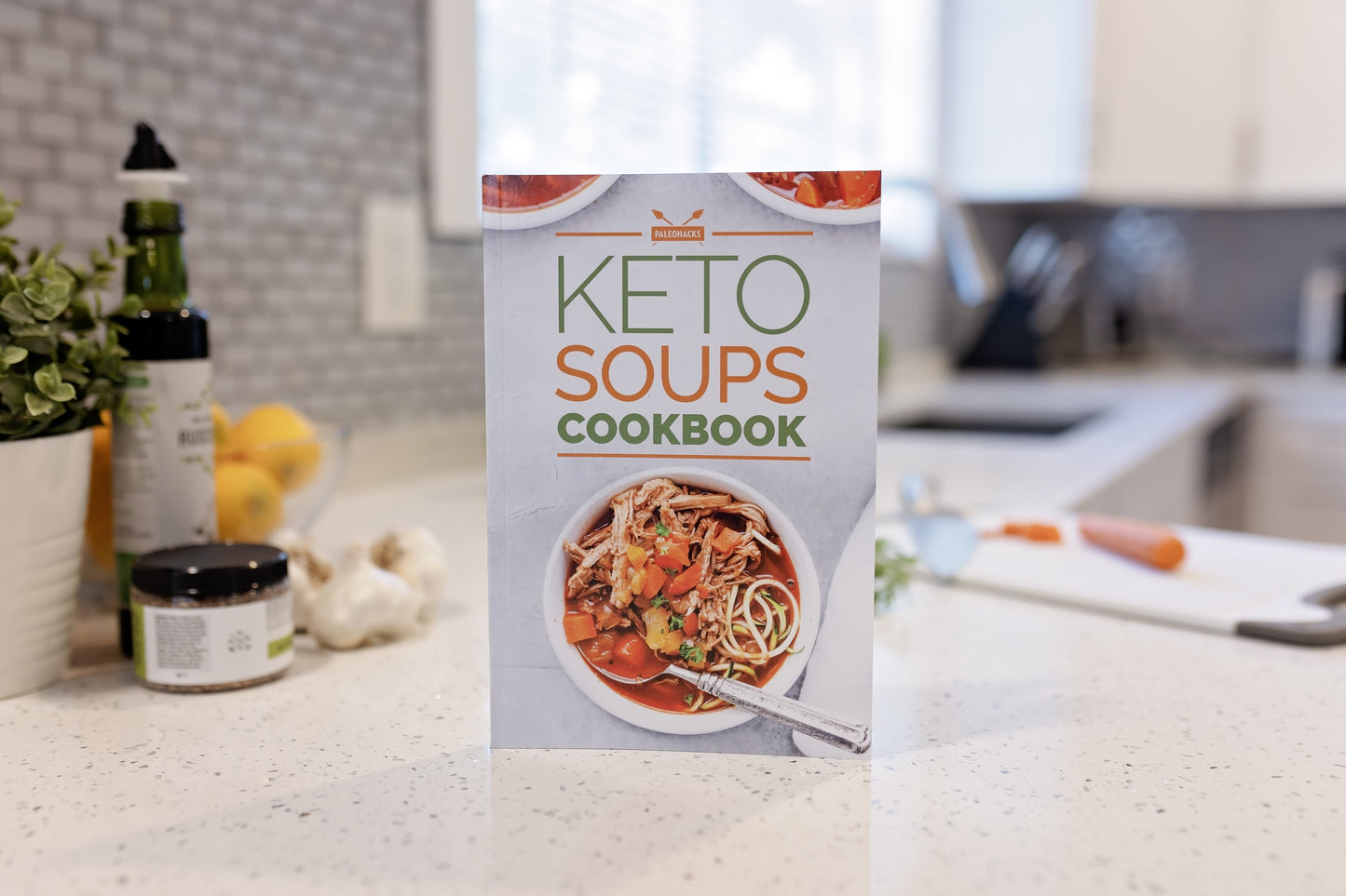 The Keto Soups Cookbook stands on a plain marble surface. Flower pots, vegetables, and some bottles can be seen.