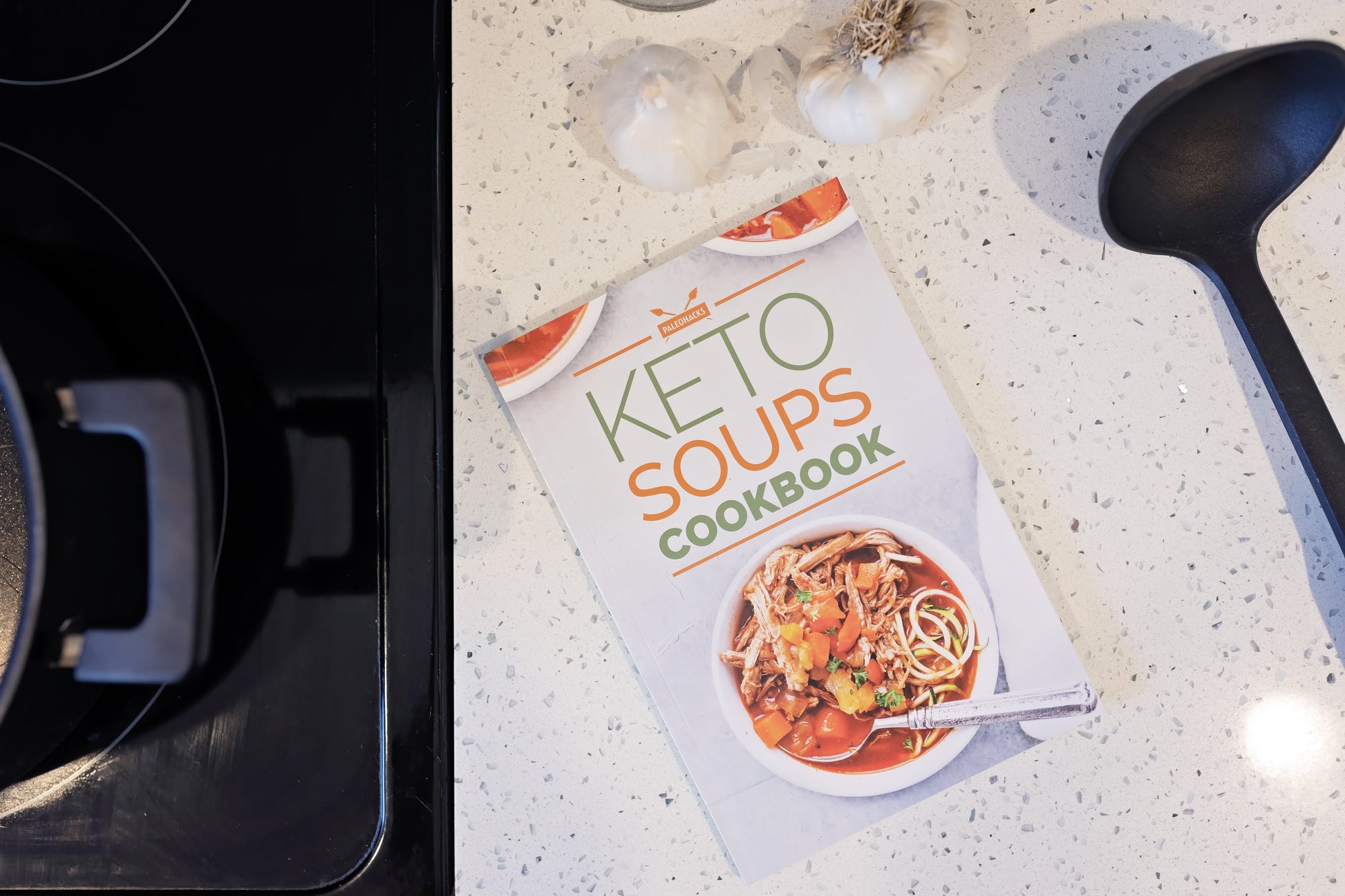 The Keto Soups Cookbook is laid on a plain marble surface, with bulbs of garlic and a spoon