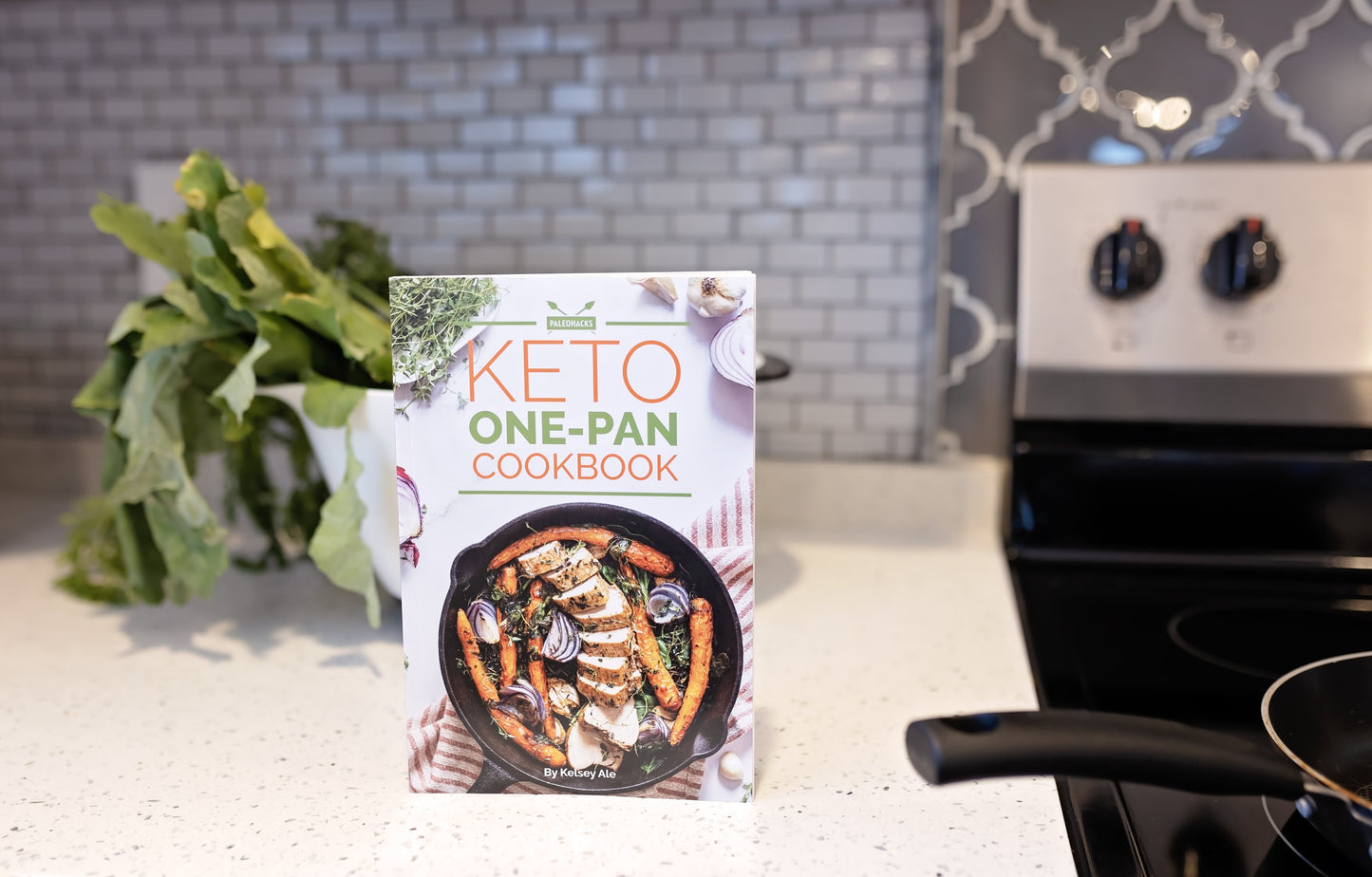 Keto One-Pan Cookbook rests on a smooth marble surface. A bowl of vegetables and a cooking appliance are visible.