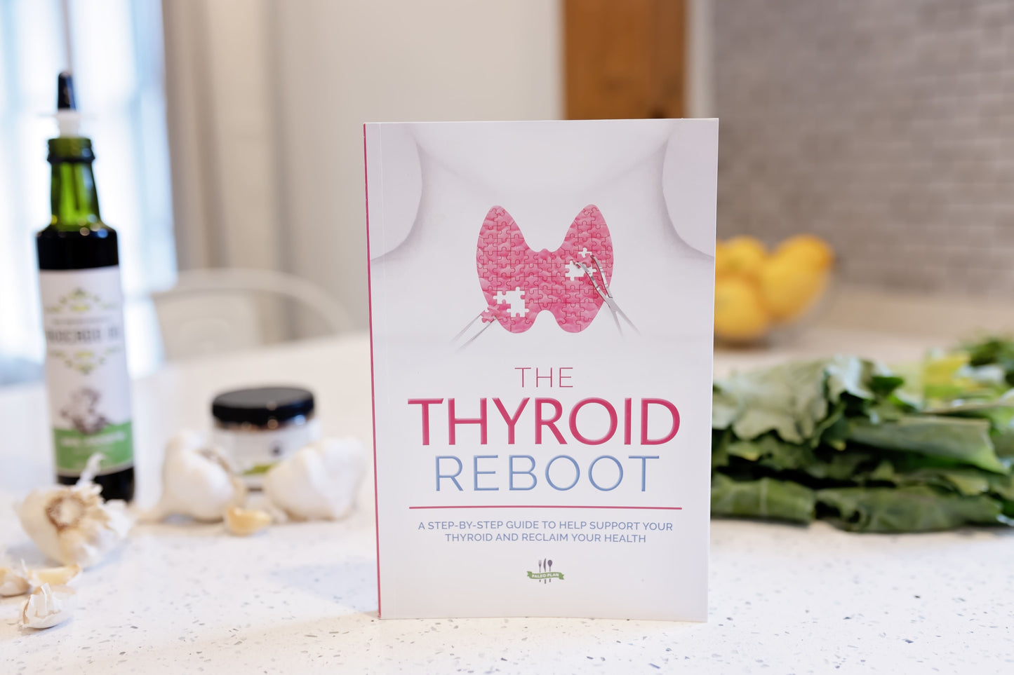 The Thyroid Reboot Book is positioned on a white plain surface, with vegetables and an avocado oil bottle in the background.