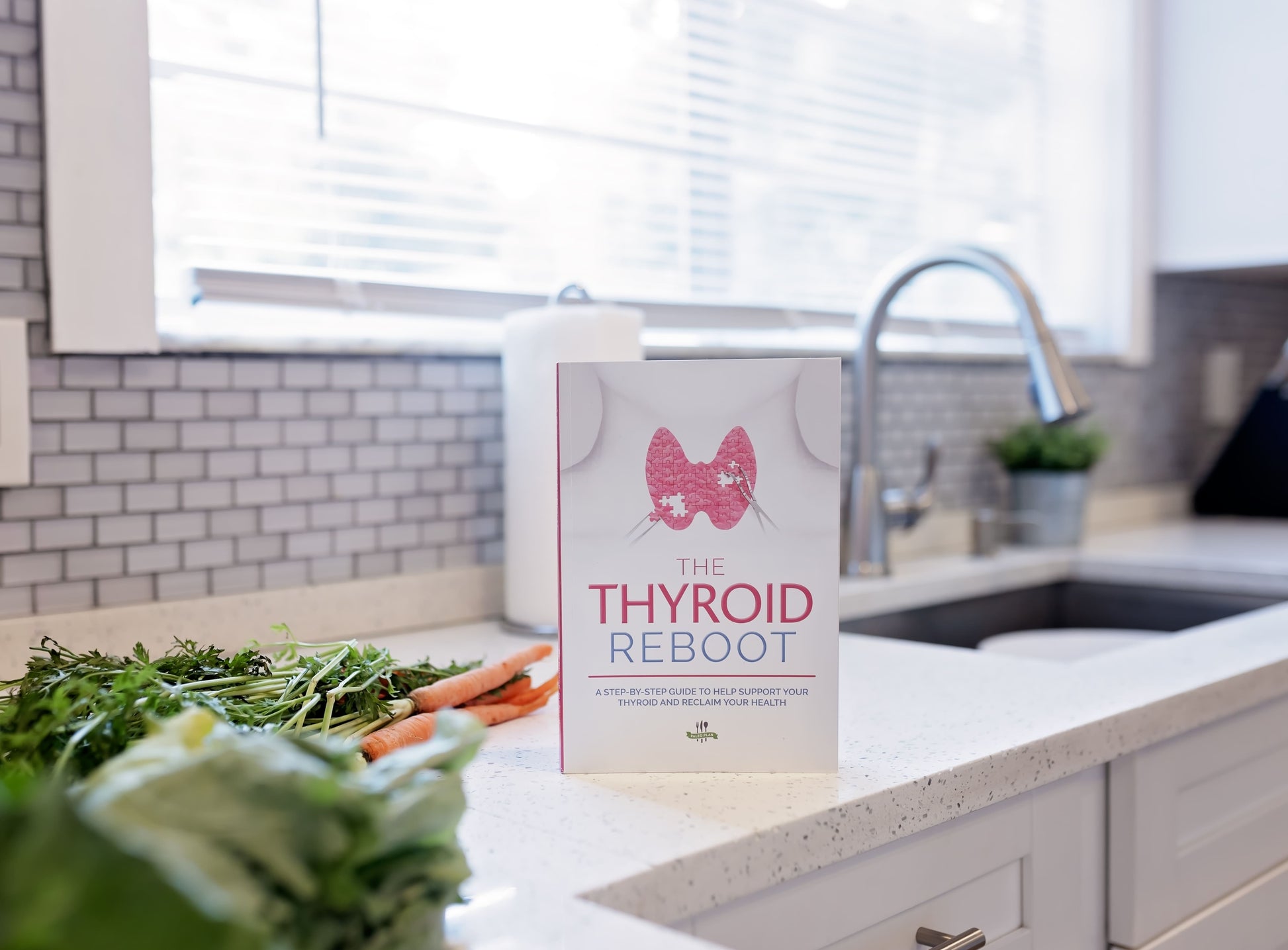 The Thyroid Reboot Book is seen standing on a white marble kitchen countertop, with vegetables in the background.