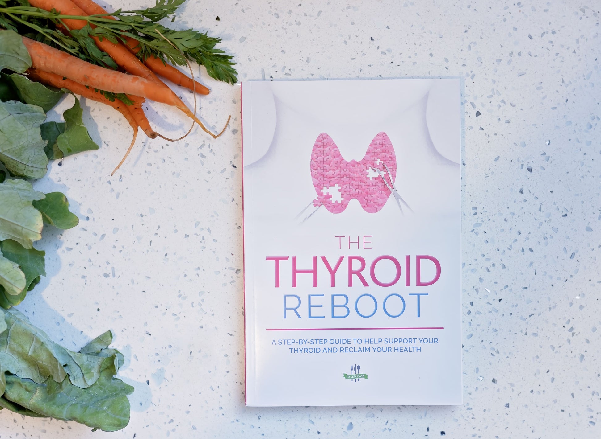 The Thyroid Reboot Book laid on a white marble surface. Vegetables can be seen on the left.