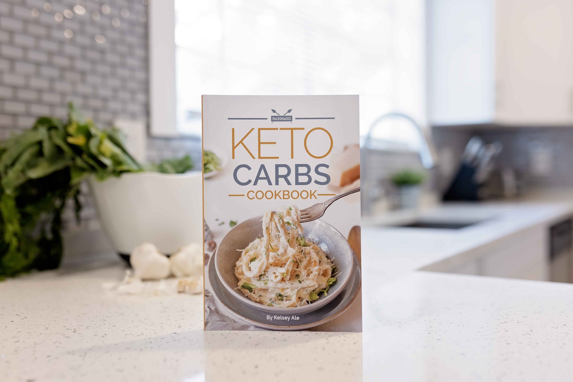 Keto Carbs Cookbook rest on a grey marble surface. A bowl of vegetables can be seen behind.