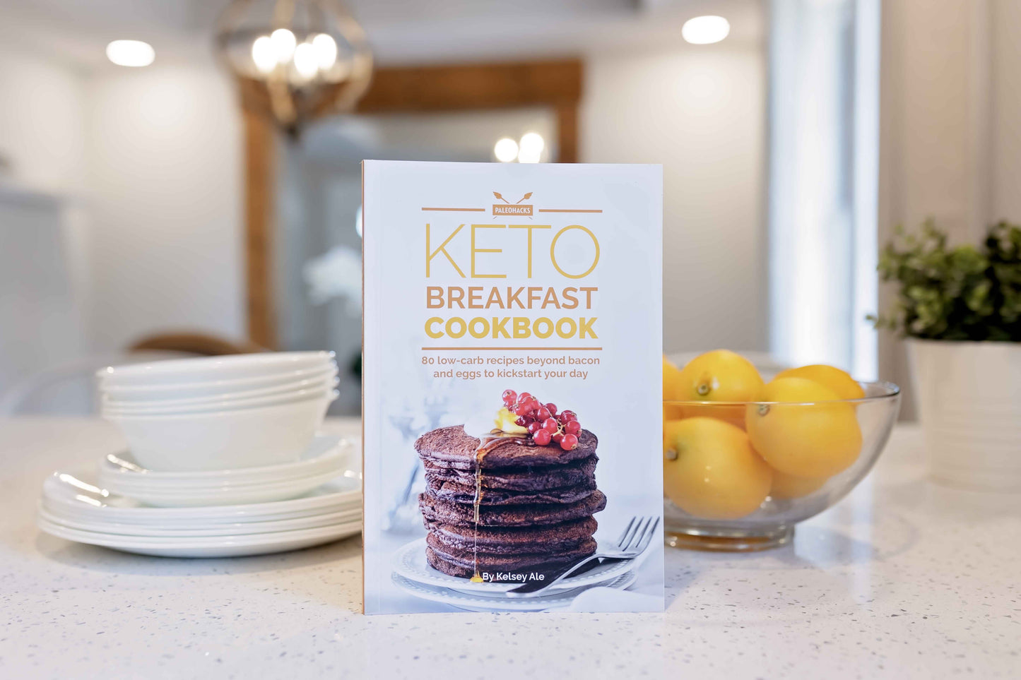 Keto breakfast cookbook is leaning against stacks of plates next to a bowl of lemons.