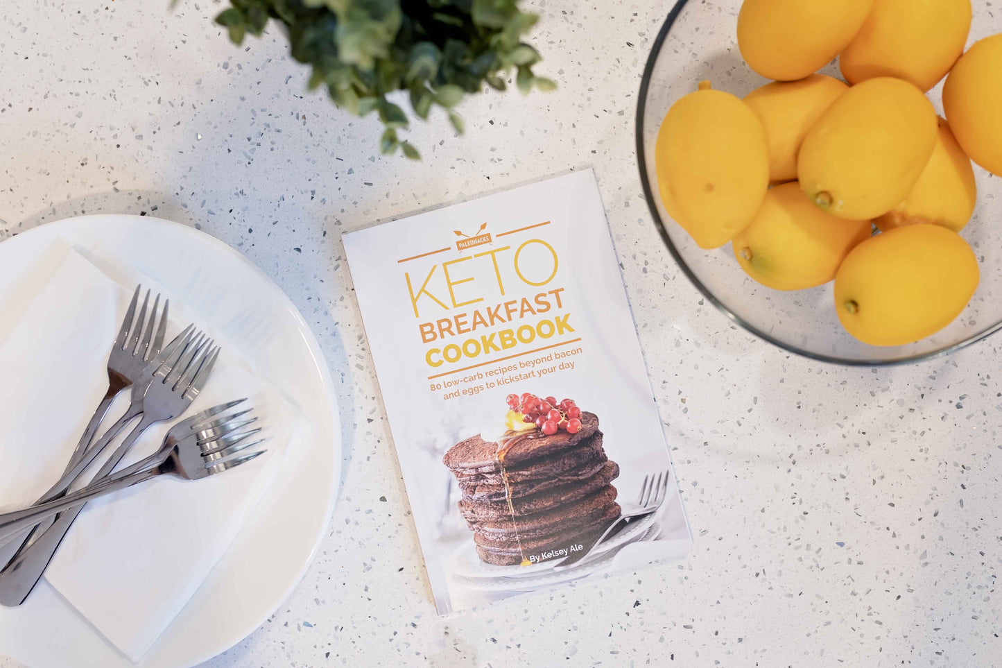 Keto breakfast cookbook laid on a marble surface with a bowl of fruits and a plate with cutlery.