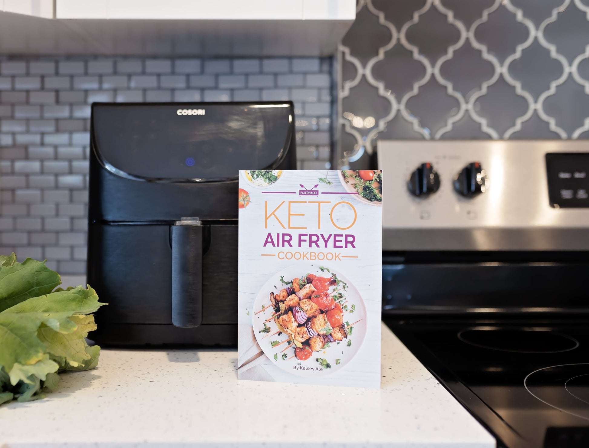 Keto air fryer cookbook leans against an air fryer and a stove.