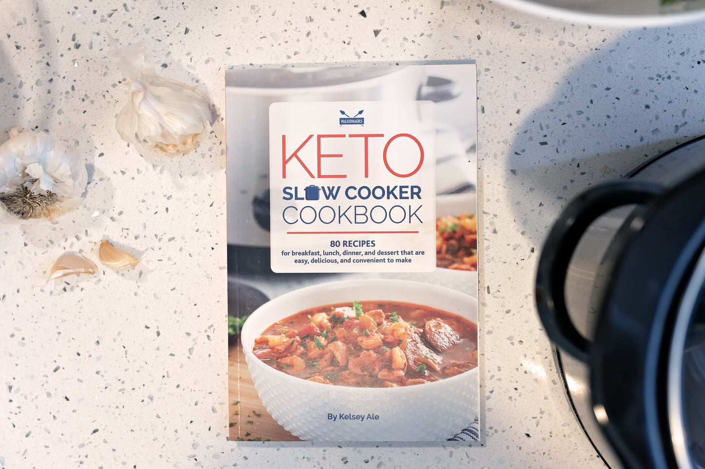 Keto slow cookbook placed on a marble surface. Garlic and cookware can also be seen in the scene