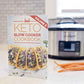 The Keto Slow Cooker Cookbook volume 2 rests on a smooth marble surface. A pot and some carrots can be seen.