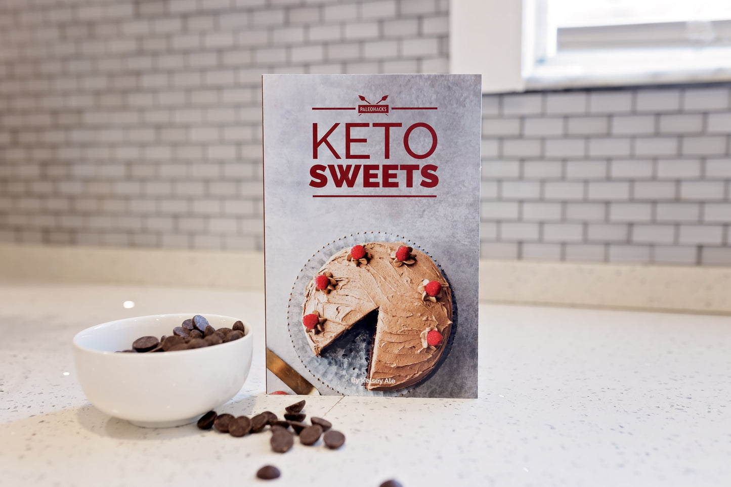 The Keto Sweets Cookbook stands on a plain white surface next to a bowl of chocolate chips.