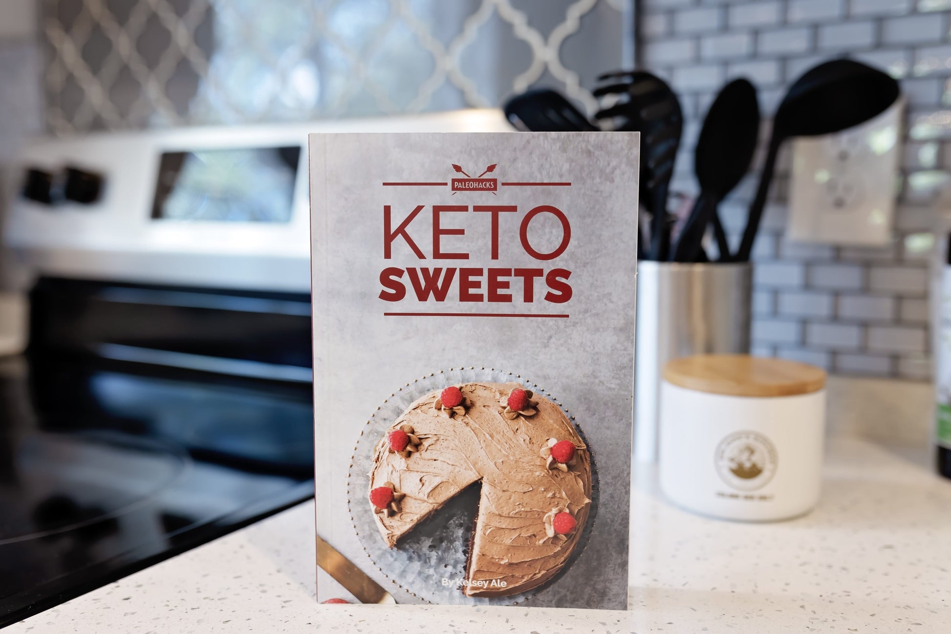 The Keto Sweets Cookbook stands on a kitchen counter. A jar of utensils can be seen.