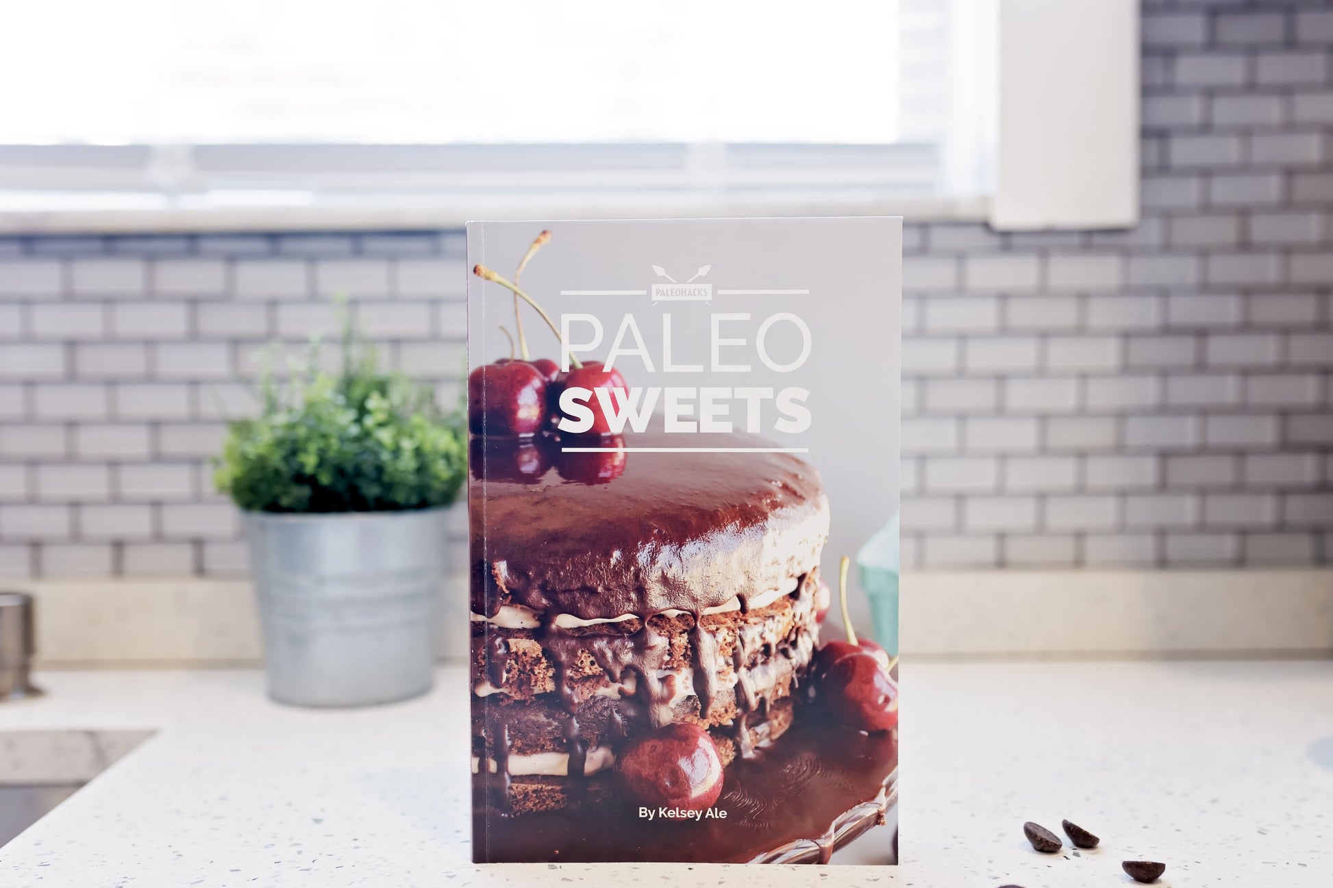 The Paleo Sweets Cookbook stands on a kitchen counter with a few chocolate chips. A flower pot can be seen in the background.
