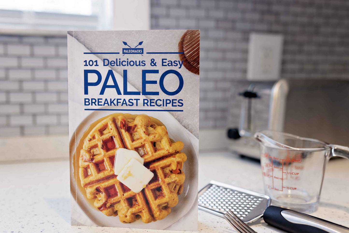 The Paleo Breakfast Recipes Cookbook stands on a kitchen counter next to a measuring cup and a grater.