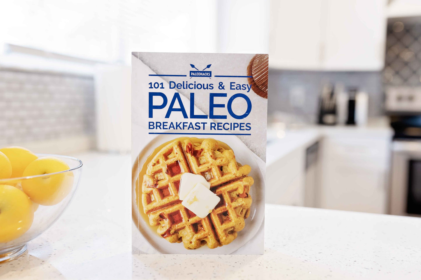 The Paleo Breakfast Cookbook stands on a kitchen counter next to a bowl of lemons.