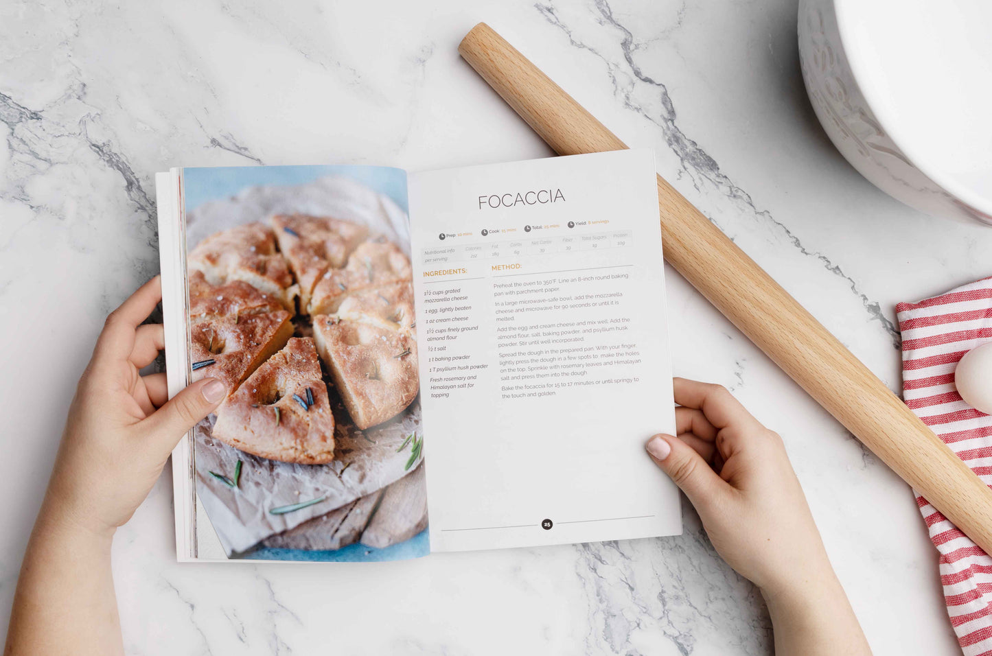 A woman holds an open keto-carb cookbook laid on a plain marble surface. A rolling pin and a kitchen cloth are also visible.