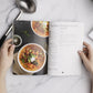 A lady opens the Keto Soup Cookbook placed on a white marble surface with a spoon.
