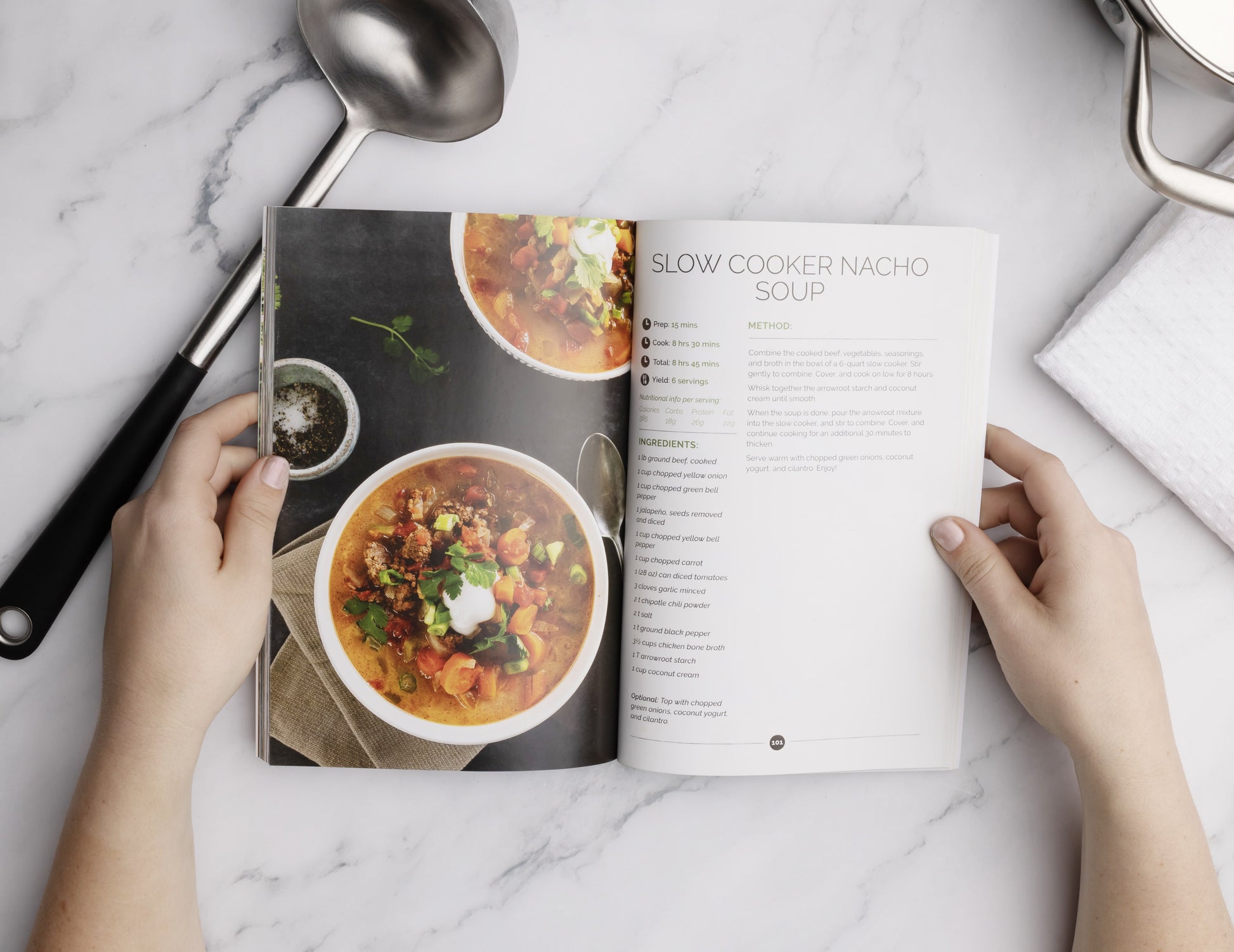 A lady opens the Keto Soup Cookbook placed on a white marble surface with a spoon.