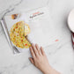 A woman's hand opens the Paleo Breakfast Cookbook on a marble surface. A plate with a whisk and a kitchen cloth are placed on the right.