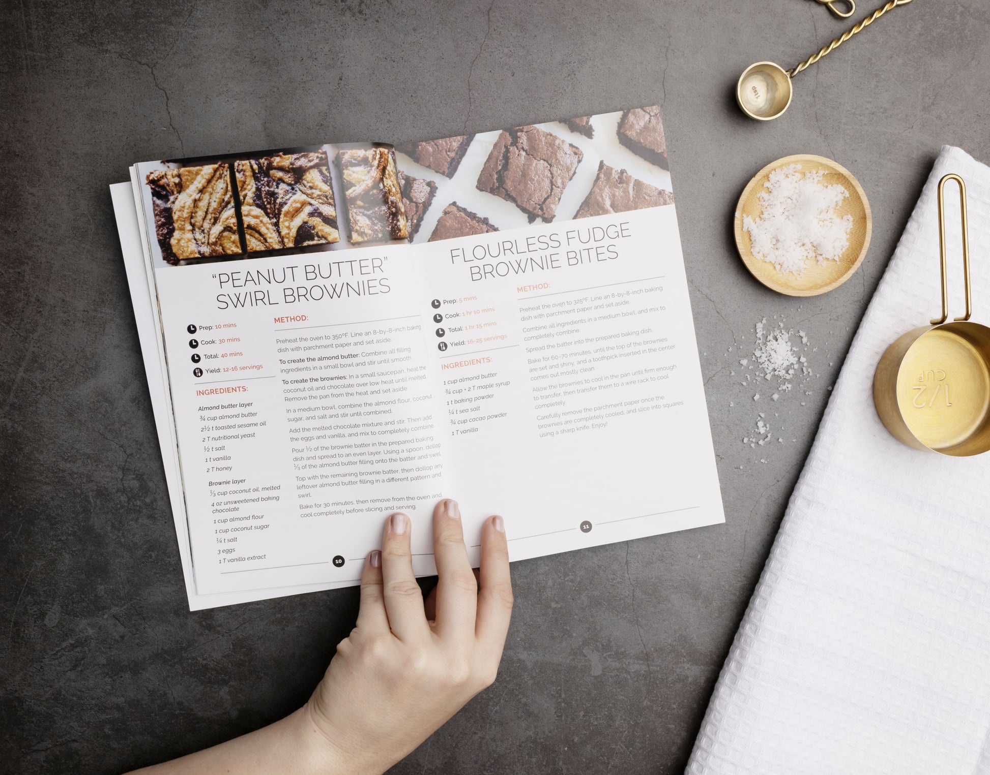 An opened Paleo Sweet Cookbook placed on a grey surface. Sea salt, measuring spoons and a kitchen cloth are seen on the right.