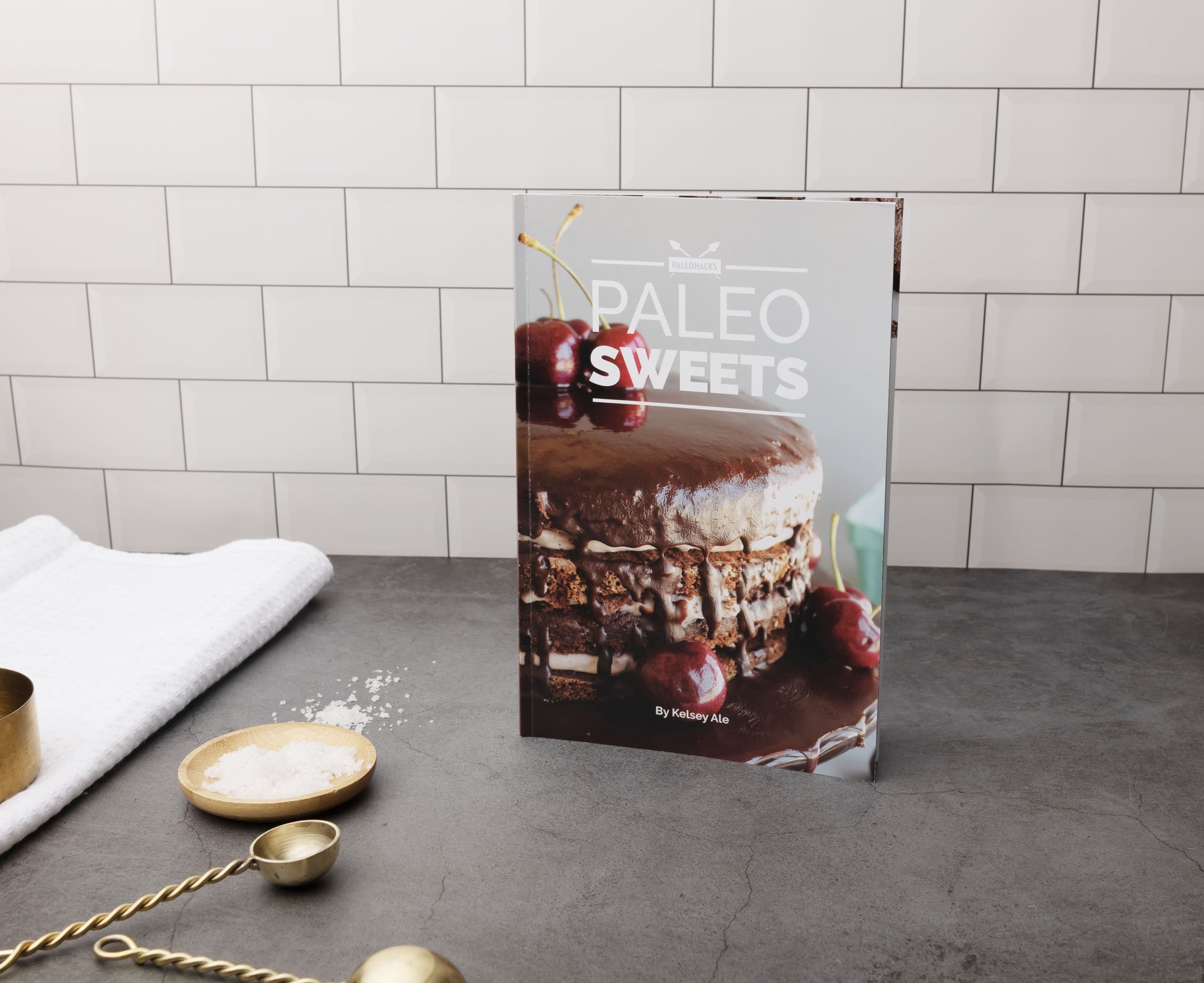 The Paleo Sweets Cookbook stands on a grey surface. Measuring spoons and a kitchen cloth can be seen on the left.