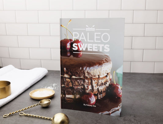 The Paleo Sweets Cookbook stands on a grey kitchen counter. Measuring spoons and a kitchen cloth can be seen on the left.