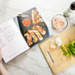 The "Everything Air Fryer" cookbook is open on a marble surface next to a wooden board displaying sliced vegetables. A bowl of vegetables and avocado oil is also visible.