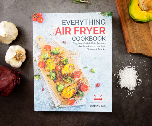 The "Everything Air Fryer" cookbook is laid out on a plain grey surface. Vegetables, cheese, and a wooden board are visible.