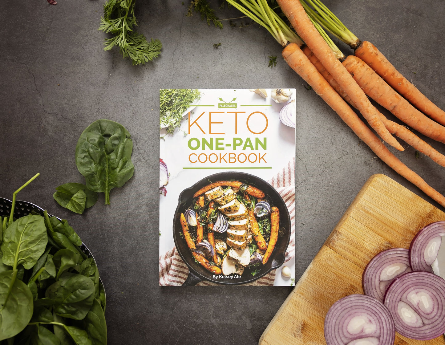Keto One-Pan Cookbook laid on a grey, plain surface, surrounded by vegetables and sliced onions.
