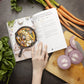 A woman's hand opens the Keto One Pan cookbook, which is laid on a grey surface. The cookbook is surrounded by vegetables and sliced onions on a chopping board.