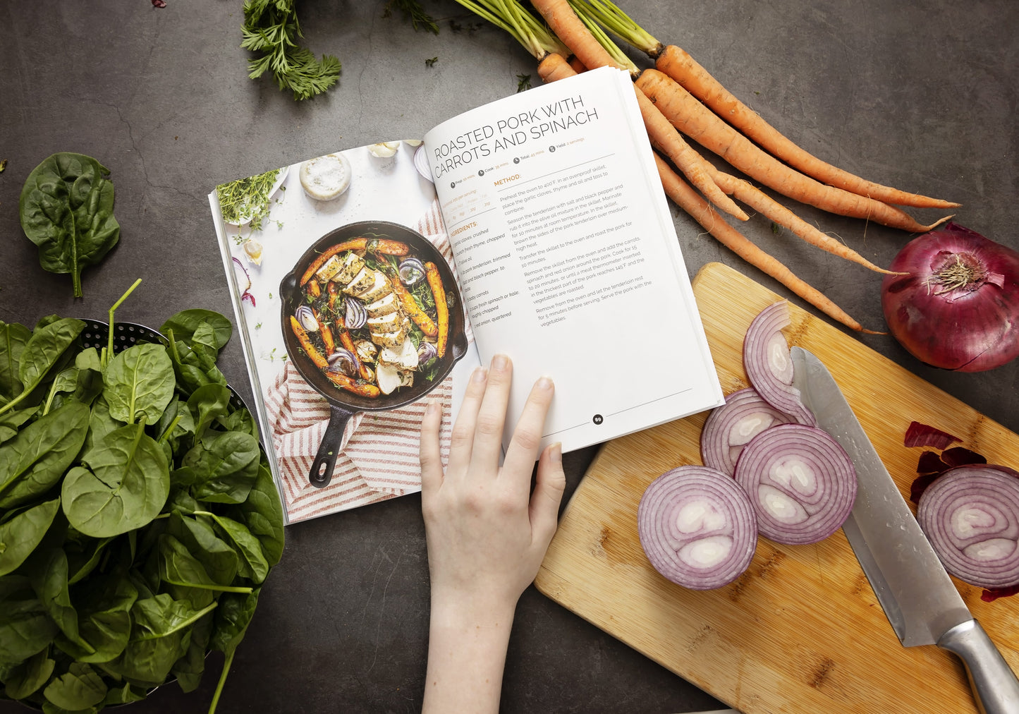A woman's hand opens the Keto One Pan cookbook, which is laid on a grey surface. The cookbook is surrounded by vegetables and sliced onions on a chopping board.