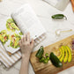 An open Thyroid Rebook cookbook laid on a kitchen cloth, placed on a plain marble surface, next to a wooden board with sliced vegetables and avocado.