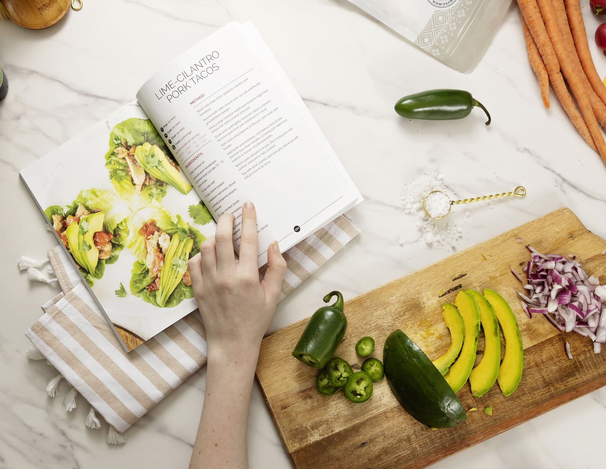 An open Thyroid Rebook cookbook laid on a kitchen cloth, placed on a plain marble surface, next to a wooden board with sliced vegetables and avocado.