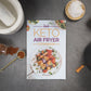 Keto Air Fryer Cookbook placed on a grey surface. Various kitchenware items, bottled herbs, and sea salt are visible alongside it.