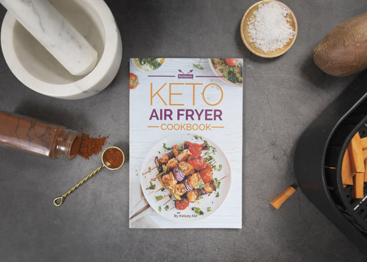 Keto Air Fryer Cookbook placed on a grey surface. Various kitchenware items, bottled herbs, and sea salt are visible alongside it.