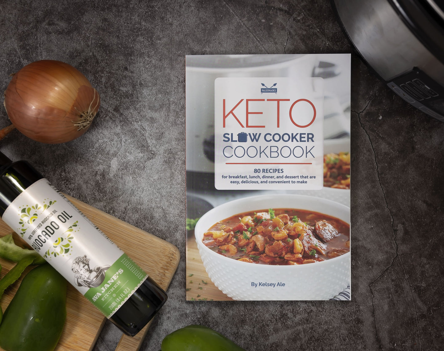 The Keto Slow Cookbook is laid on a plain grey surface. Avocado oil and vegetables can be seen on a wooden board.