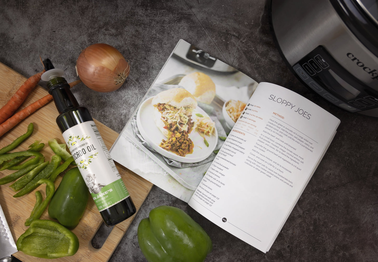  An opened Keto slow cookbook placed on a grey surface, with a wooden board having vegetables and a bottle of avocado oil.