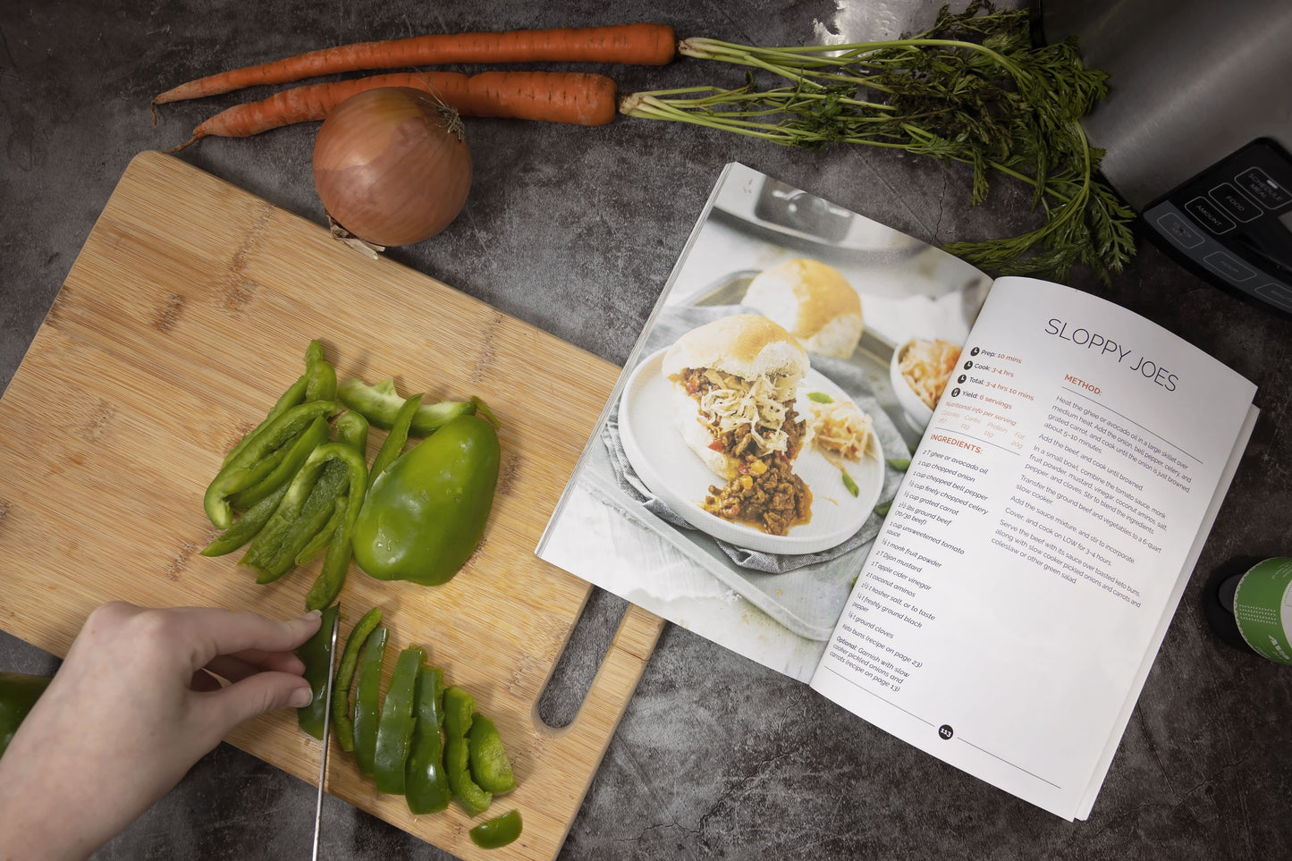Vegetables are sliced on a wooden board placed on a plain grey surface with an opened Keto slow cookbook resting on it. Carrot and onion are visible.