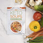 The Keto Soups Cookbook placed on a marble plain surface with some vegetables by the right.