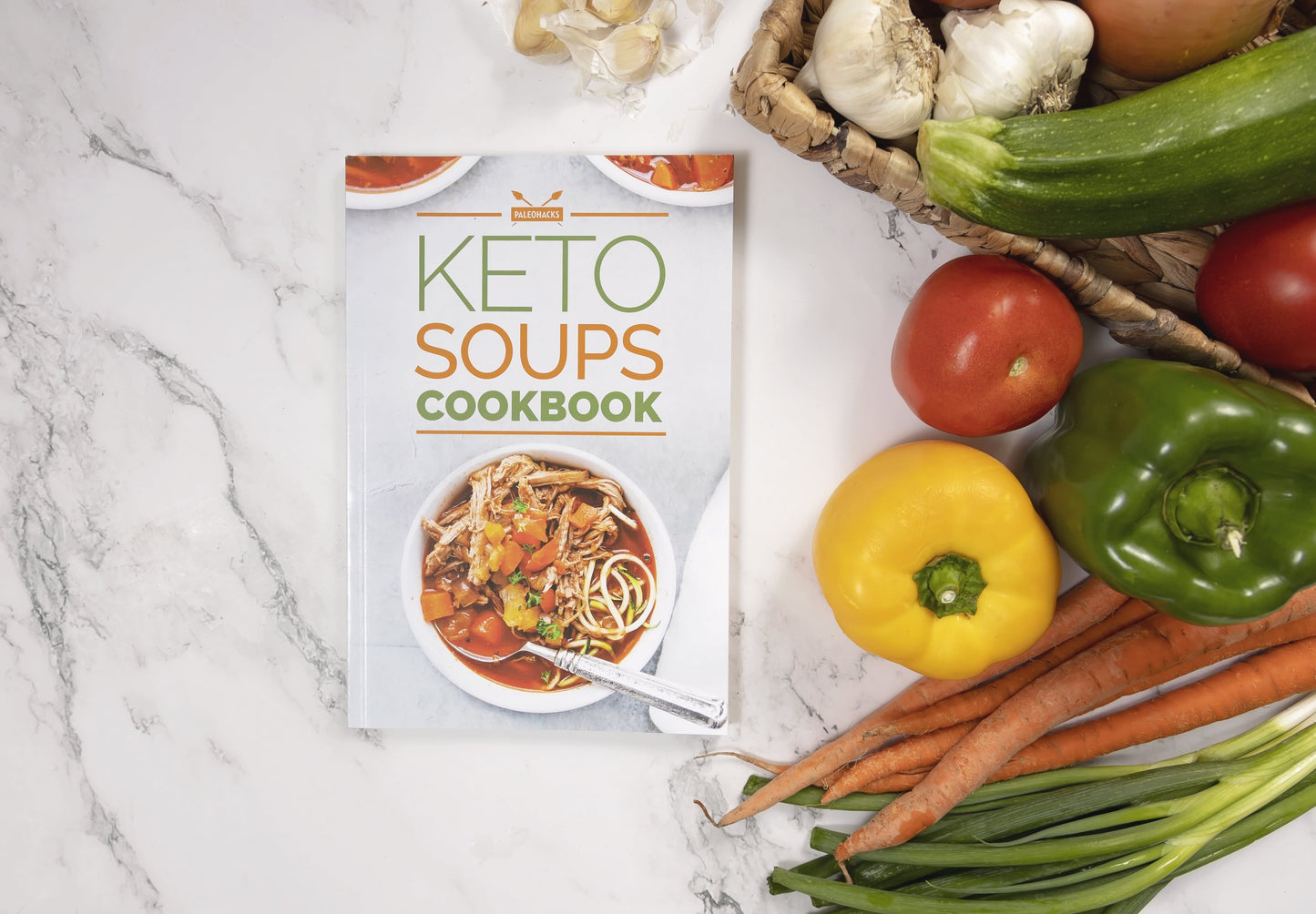 The Keto Soups Cookbook placed on a marble plain surface with some vegetables by the right.