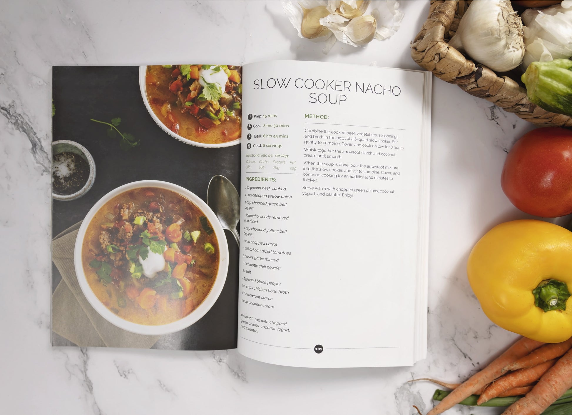 An opened Keto Soups Cookbook placed on a white marble surface with vegetables on the right.