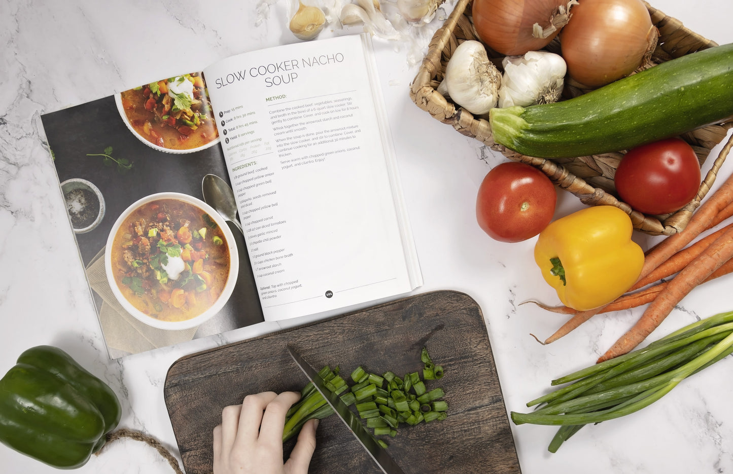 A lady's hand seen dicing spring onions on a black cutting board next to an opened Keto Soup Cookbook on a white marble surface, with vegetables on the right.