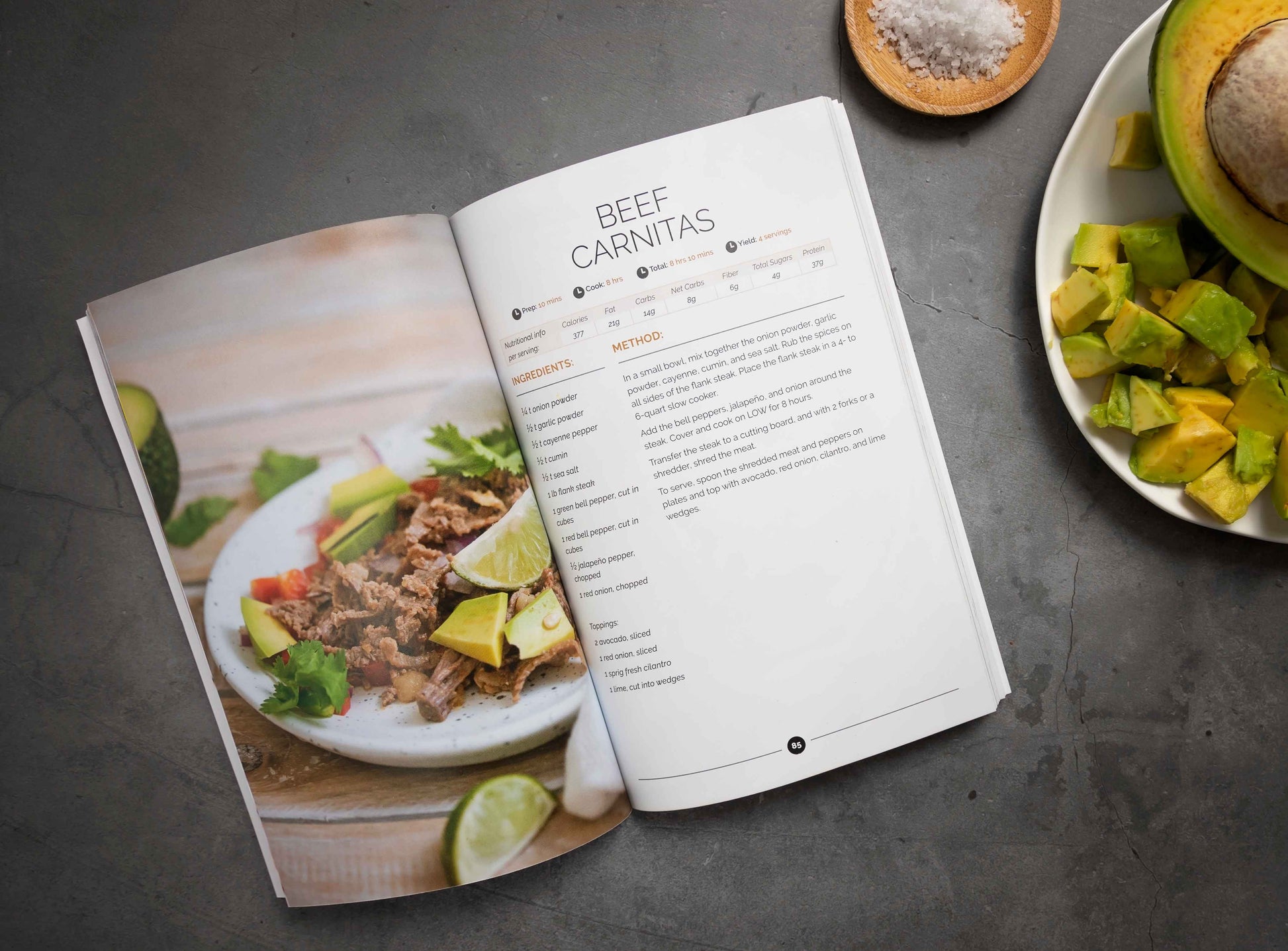 An open Keto Slow Cooker Cookbook volume 2 is placed on a plain grey surface. A plate of avocado and a bowl of salt are seen.