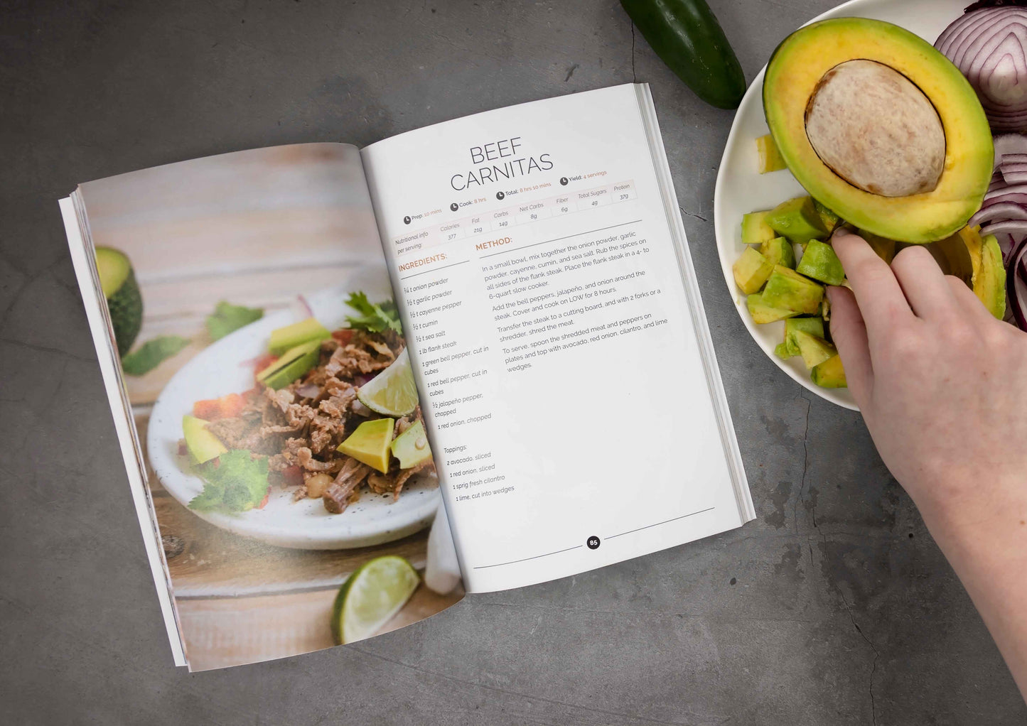 An open Keto Slow Cooker Cookbook -Volume 2, is laid on a grey surface. A lady's hand can be seen picking avocados from a plate of avocados and onions.