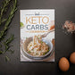 Keto Carbs Cookbook laid on a plain grey surface, with rosemary, sea salt, and eggs.