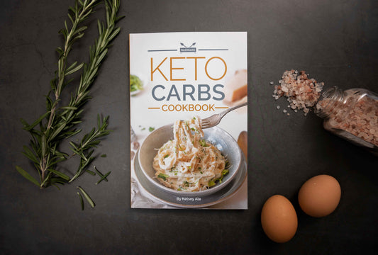 Keto Carbs Cookbook laid on a plain grey surface, with rosemary, sea salt, and eggs.