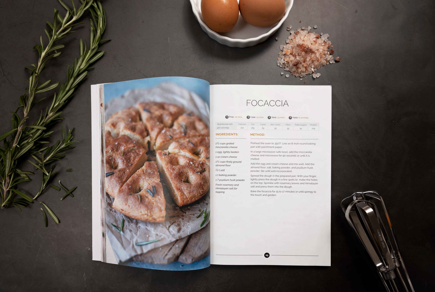 An opened keto carbs cookbook placed on a grey surface. There is rosemary and sea salt nearby. A white bowl of eggs, and a mixer is also visible.