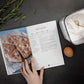 A woman's hand opens the keto carbs cookbook on a grey surface. A bowl of flour in the mixed, an egg and a sprig of rosemary can be seen.